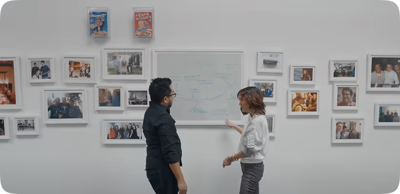Two people standing and discussing in front of a whiteboard surrounded by framed photographs on a white wall.