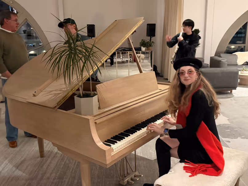 Woman in black outfit and red scarf playing a light wooden grand piano indoors with three people in the background.