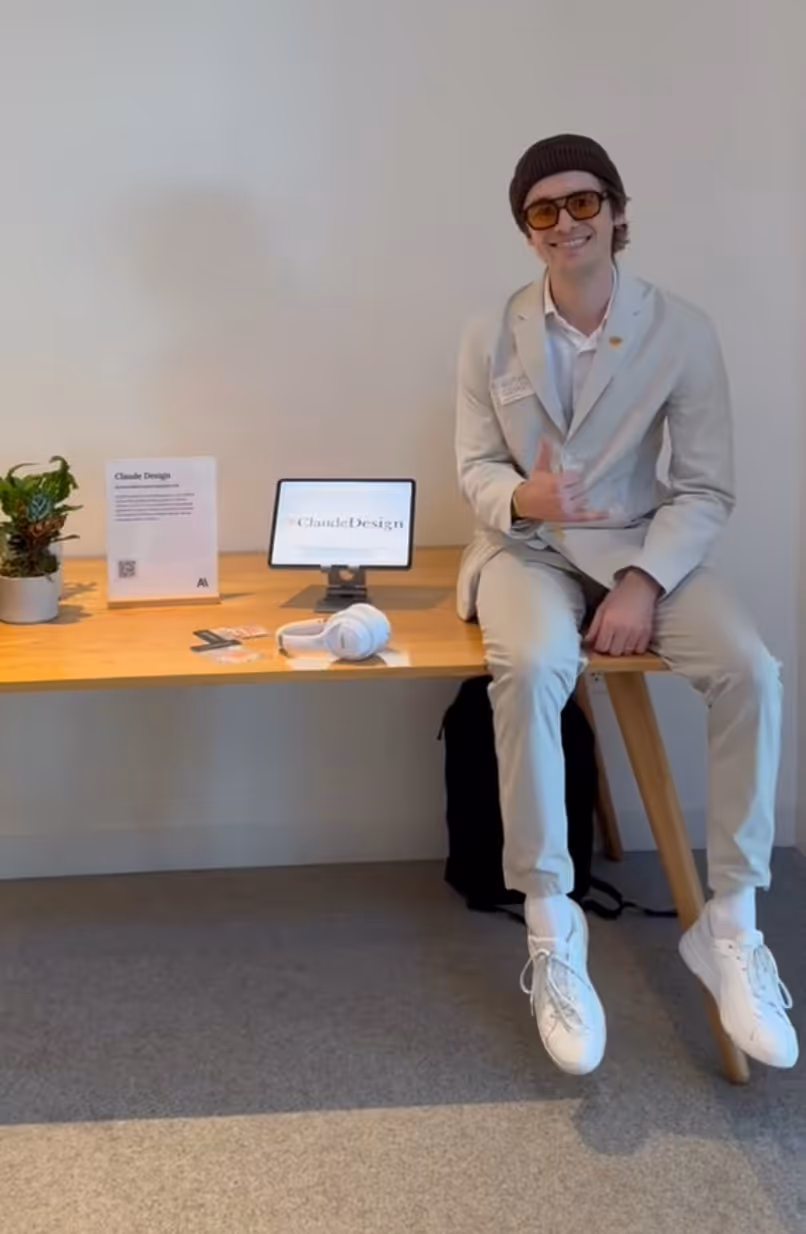 Man in light gray suit and beanie sitting on wooden table with headphones, plant, and tablet displaying Claude Design logo.