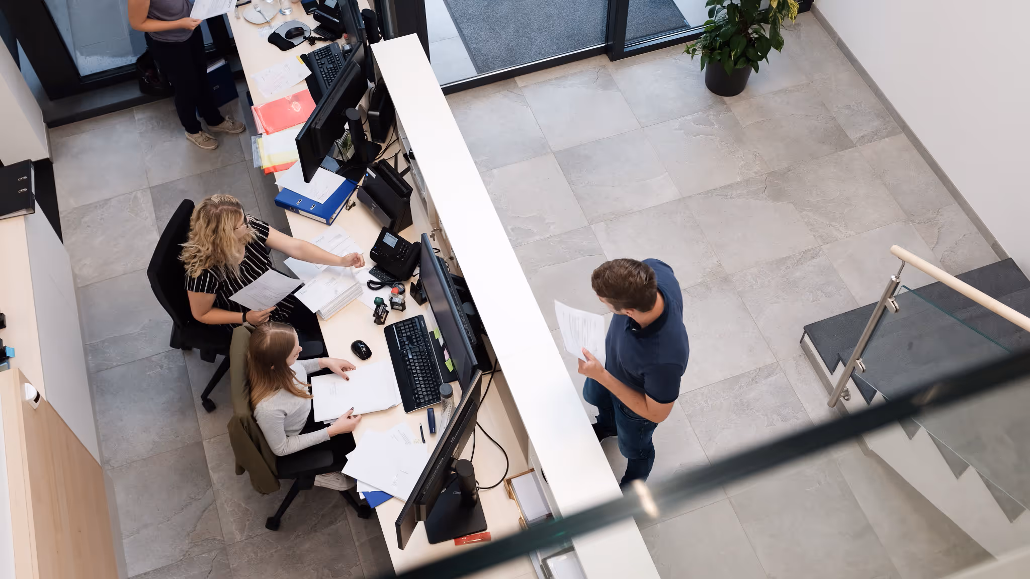 Bürosituation mit zwei Frauen an einem Schreibtisch und einem Mann, der mit Papieren vor dem Empfangstresen steht.