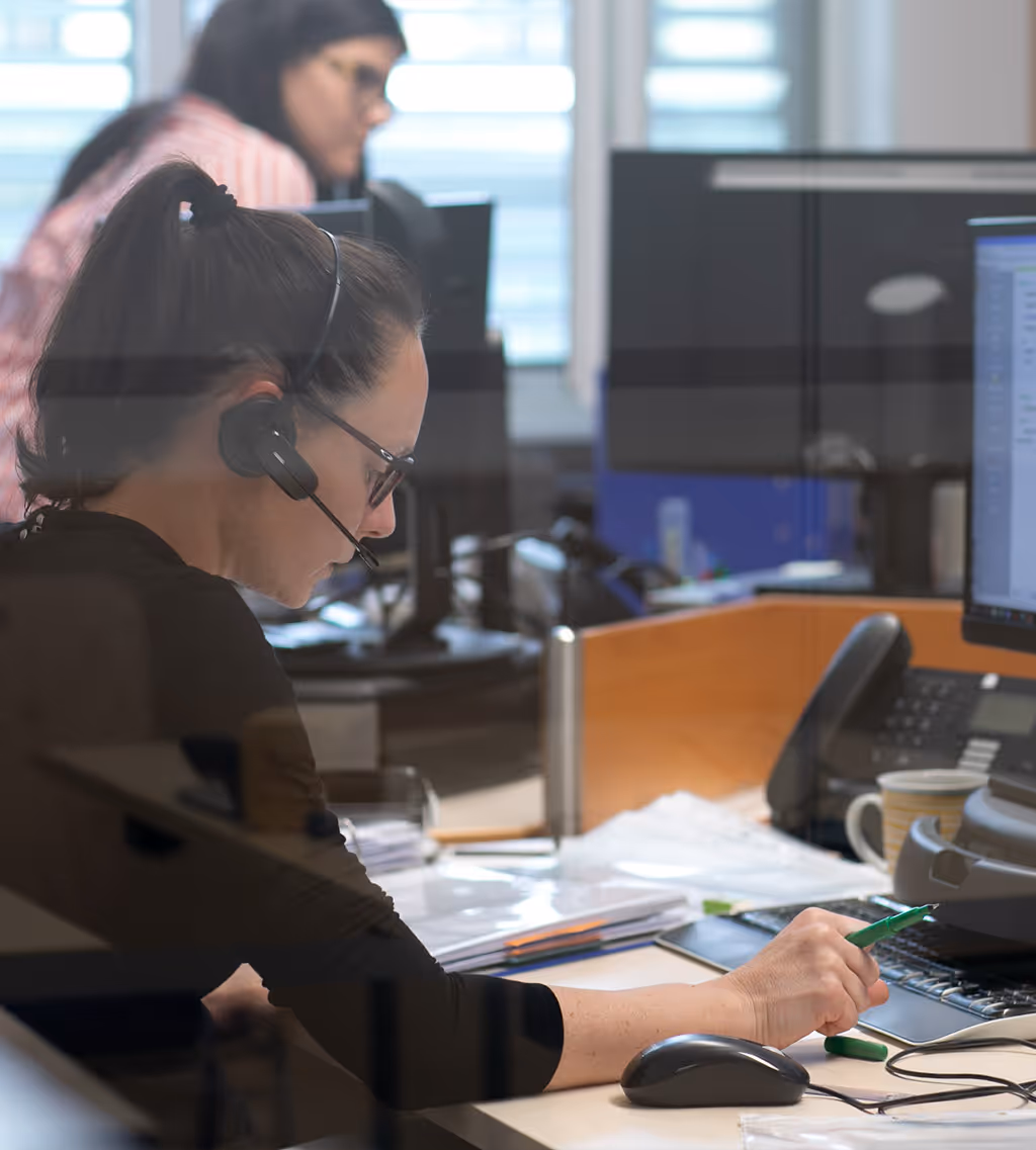 Frau mit Headset arbeitet konzentriert an einem Schreibtisch in einem Büro mit Computer und Telefon.