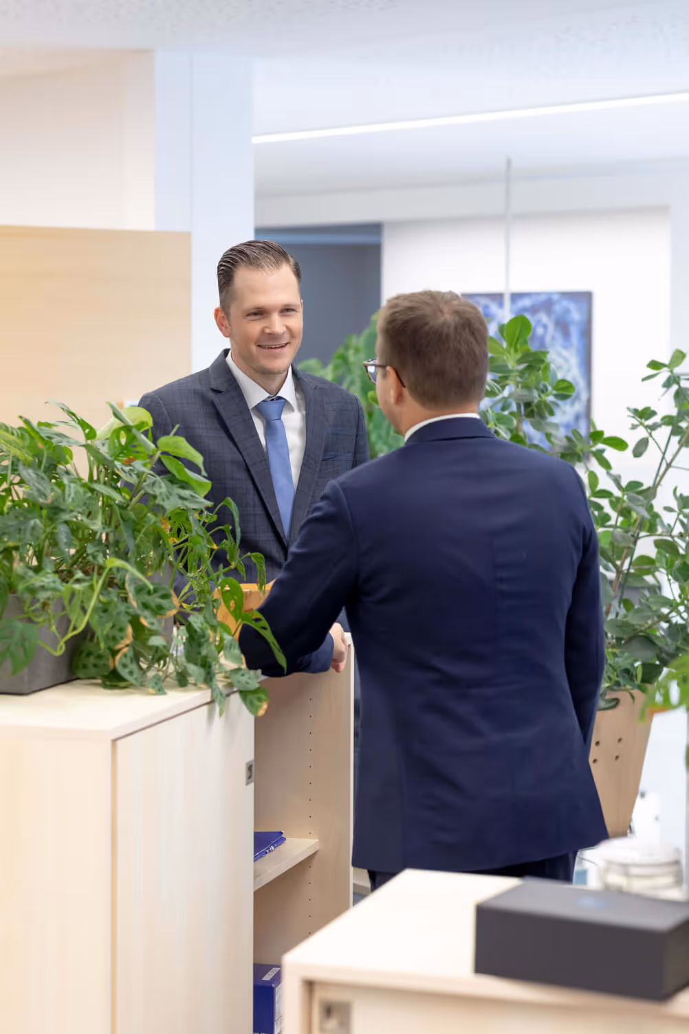 Zwei Männer in Anzügen schütteln sich in einem hellen Bürogebäude mit Pflanzen die Hand.