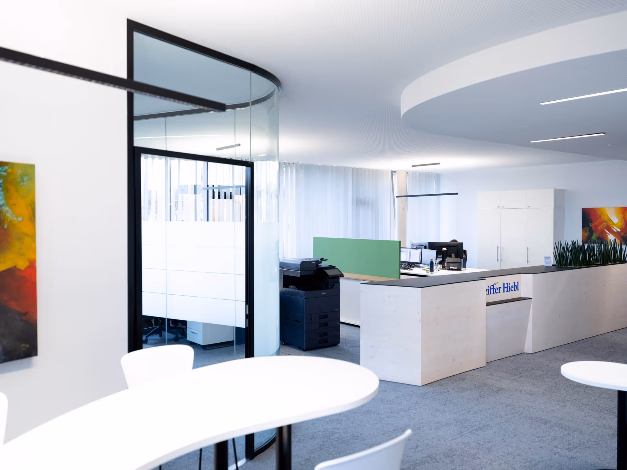 Moderner Bürobereich mit weißen Möbeln, Glaswand, Schreibtischen mit Computern und Pflanzen.