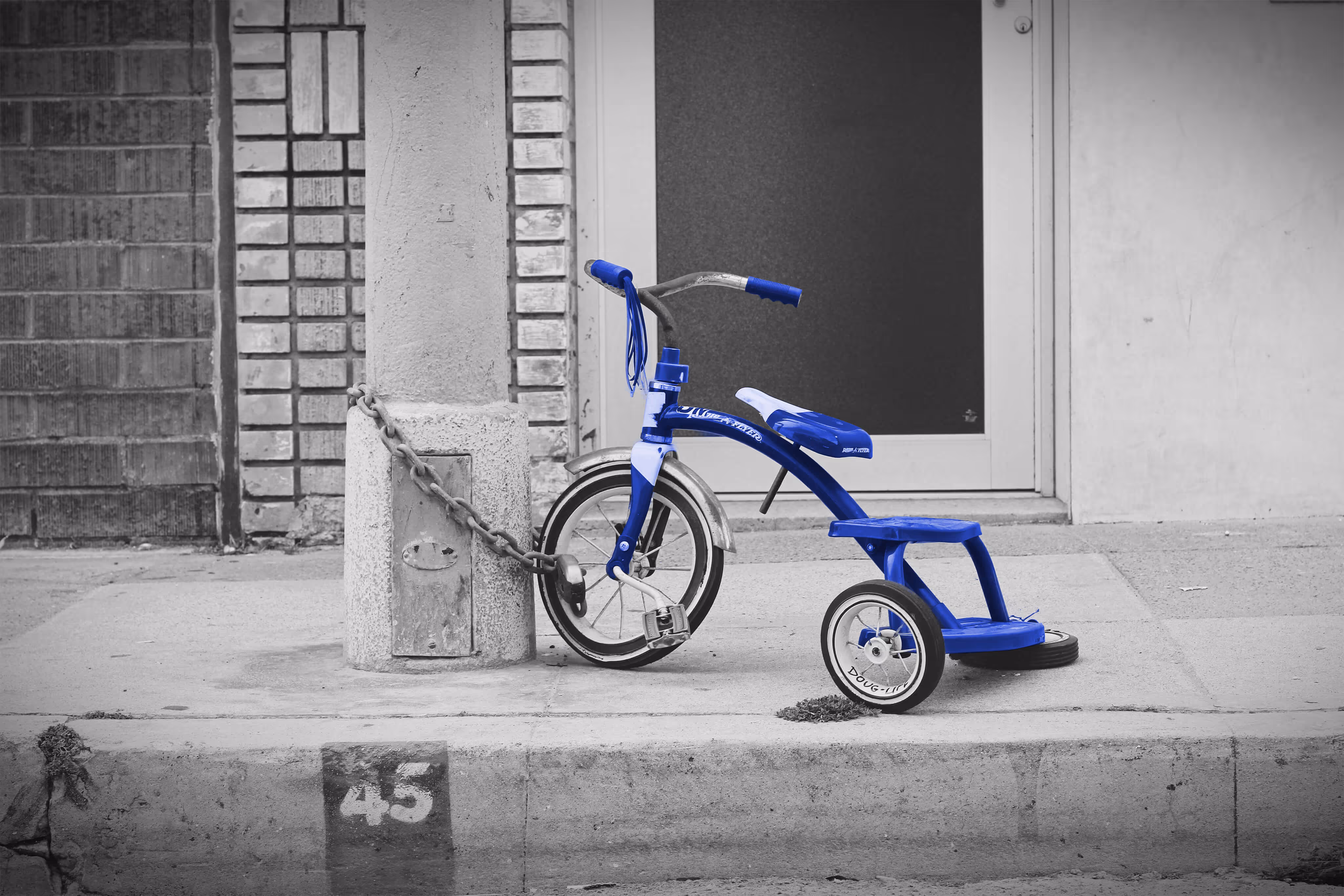 Blau lackiertes Dreirad mit Pedalen, angekettet an einen Betonpfosten vor einem Gebäude.