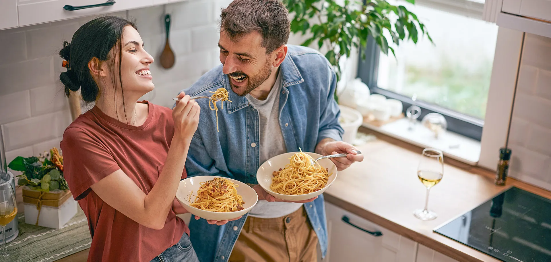 Eine Frau und ein Mann stehen in einer Küche, lachen und essen Spaghetti