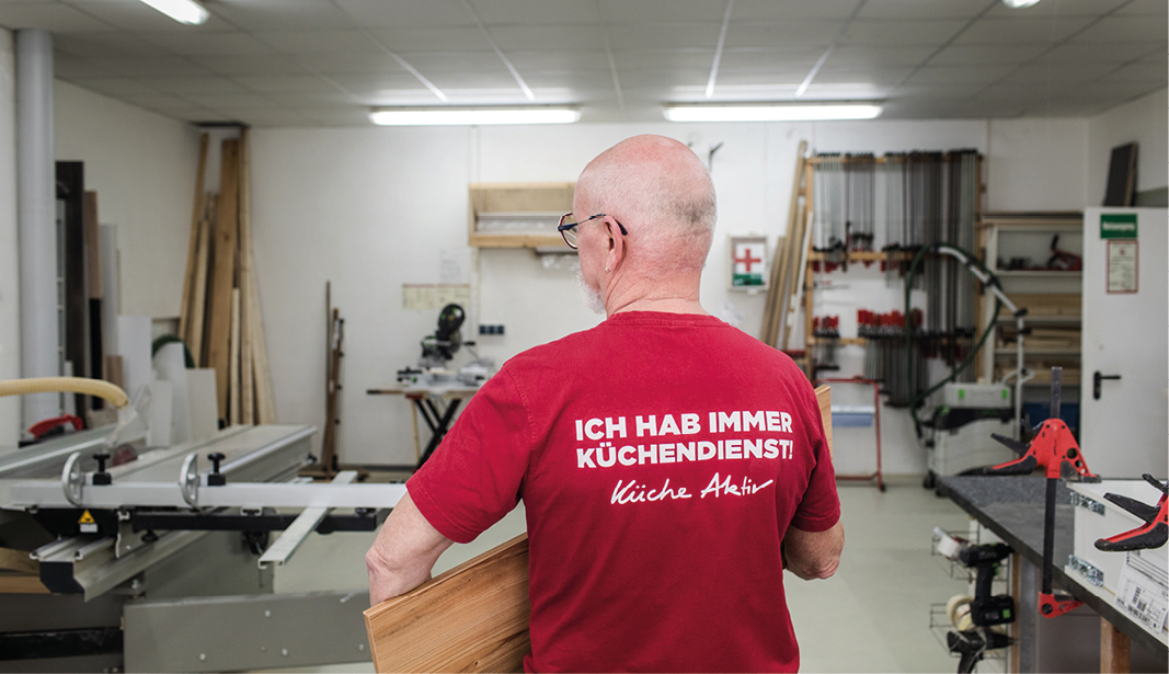 Moderner Küchenschreiner bei der Arbeit in der Werkstatt mit rotem T-Shirt mit Aufdruck „Ich hab immer Küchendienst!“ – handgefertigte Küchen von Küche Aktiv, hochwertige Maßanfertigung und individuelle Küchenmontage.