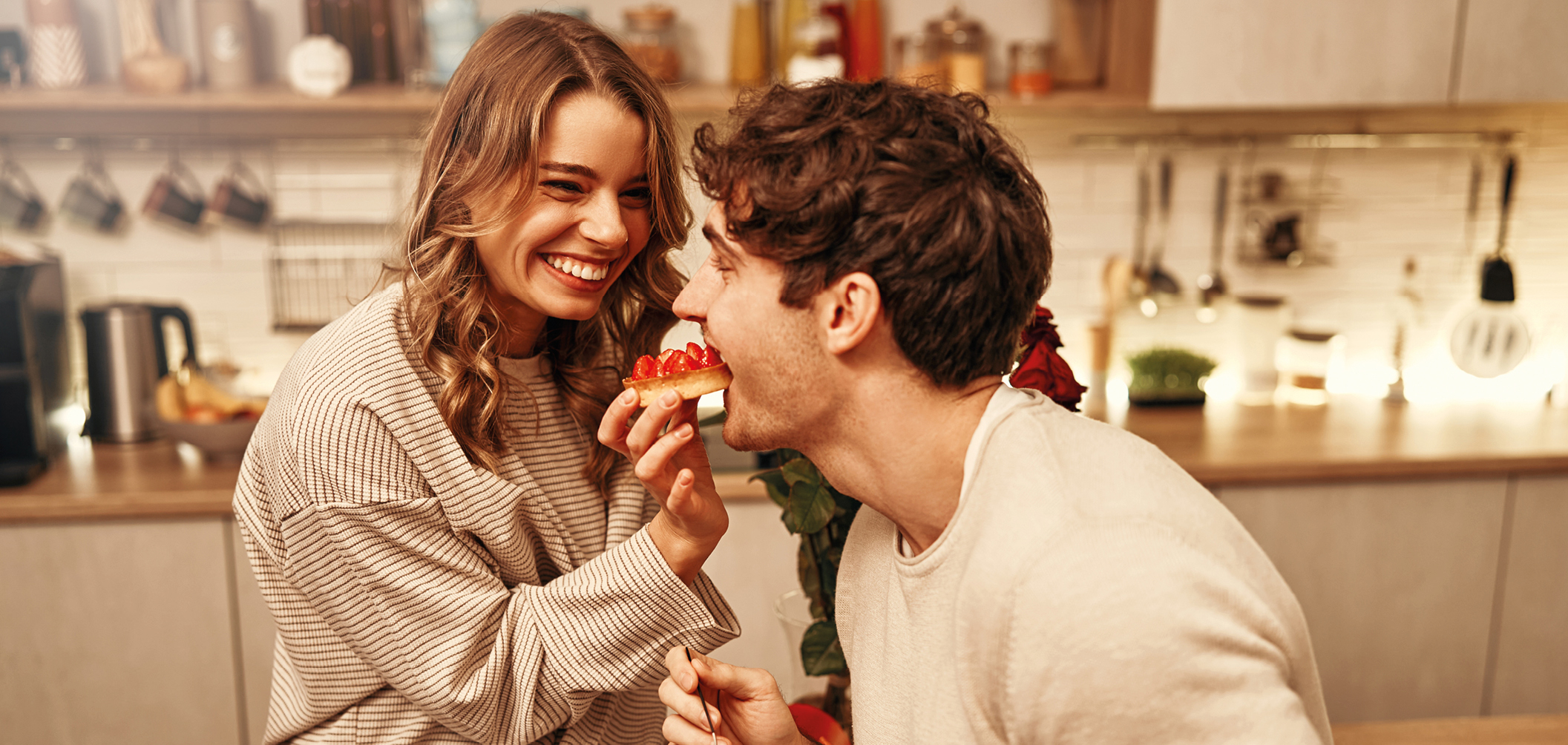 Junges Paar lacht und teilt einen Erdbeerkuchen in einer modernen Küche mit warmem Licht und Holzdekor. Symbol für Genuss, Liebe und gemütliche Momente zuhause in stilvoller Kücheneinrichtung.