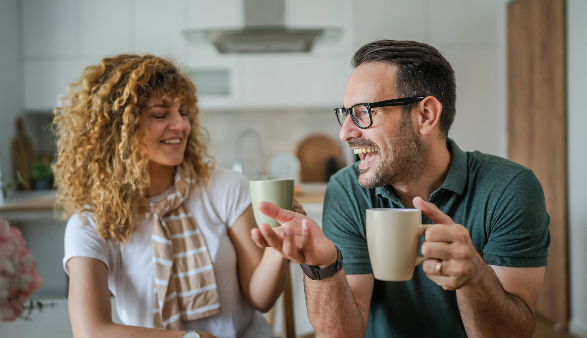 Ein lachendes Paar genießt gemeinsam Kaffee in einer modernen Küche – harmonischer Moment bei Tageslicht, der Gemütlichkeit, Kommunikation und Wohnqualität im modernen Küchenumfeld vermittelt.