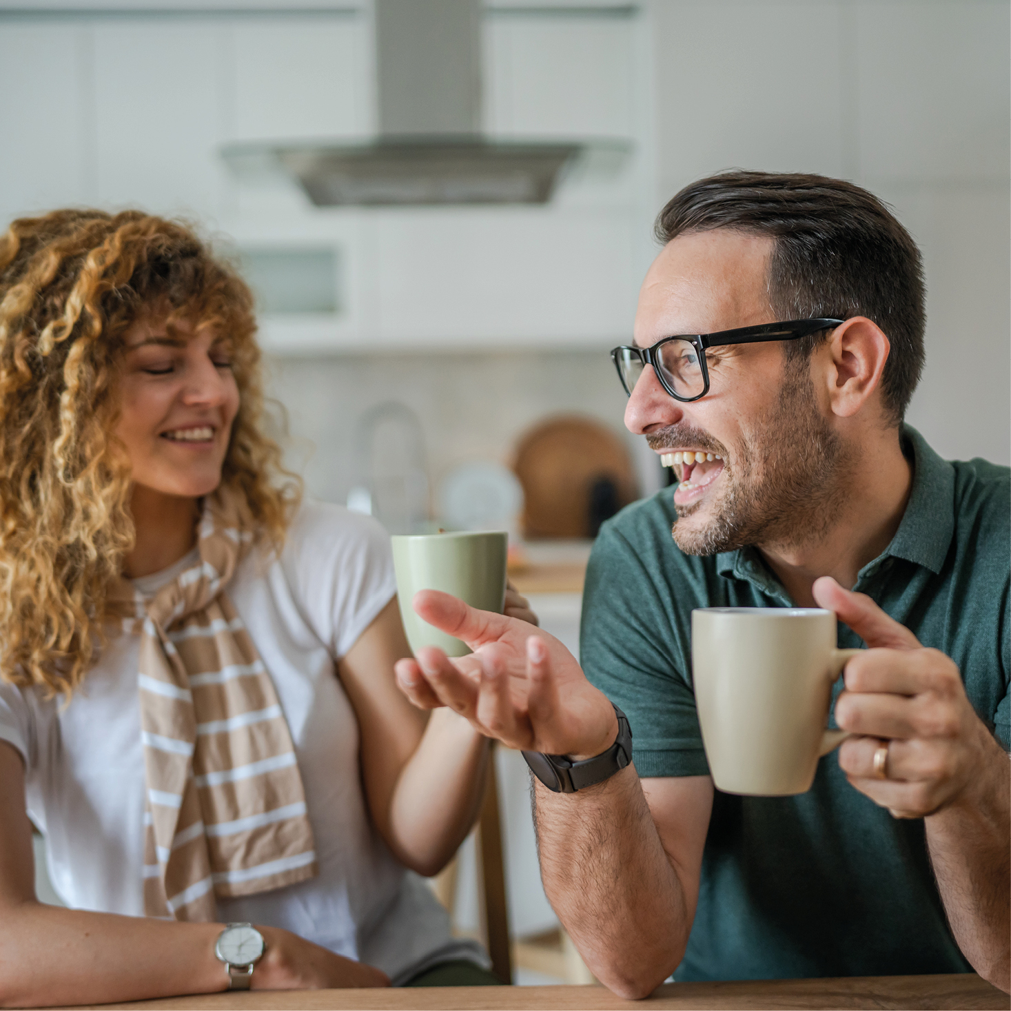 Junges Paar lacht gemeinsam bei einer Tasse Kaffee in heller Küche – entspannte Atmosphäre mit Fokus auf Kommunikation und Wohlfühlen. Symbol für Genussmomente und gemütliche Gespräche im Zuhause.
