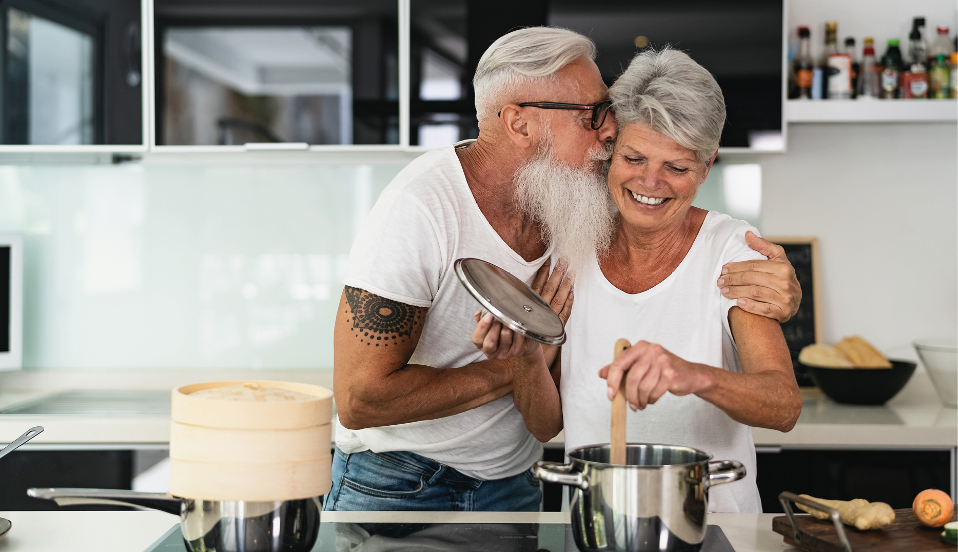 Älteres Paar kocht gemeinsam in moderner Küche und lacht miteinander – Mann küsst Frau auf die Wange, während sie am Herd rührt. Symbol für Liebe, Genuss und gemeinsames Kochen im Alltag.