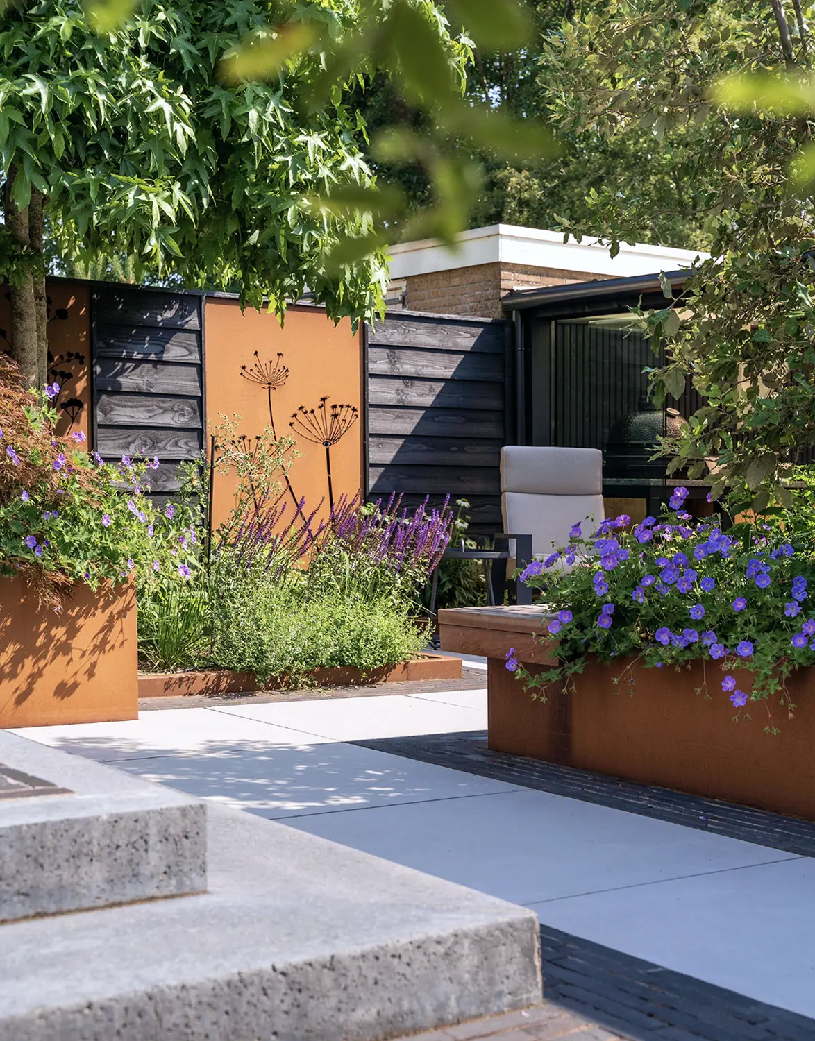 Moderne tuin met betonnen paden, metalen plantenbakken met paarse bloemen, groene struiken en een grijze tuinstoel.