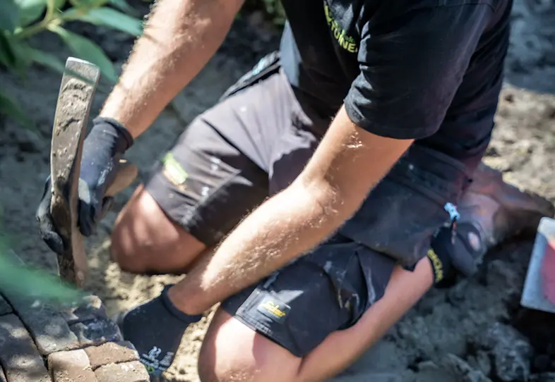 Persoon knielt op de grond en gebruikt een gereedschap om stenen of tegels te leggen in de tuin.