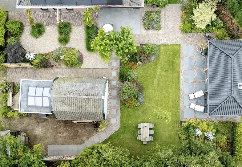Luchtfoto van een tuin met twee woningen, grind- en betegelde paden, groene grasvelden, bloembedden, en een houten tuinset.