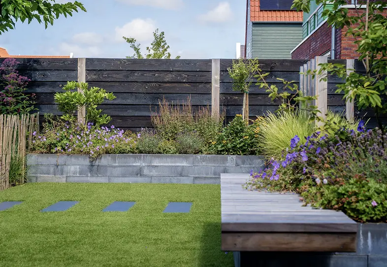 Moderne tuin met een grasveld, grote plantenbakken met bloeiende en groene planten, houten schutting en een houten bank aan de rechterkant.