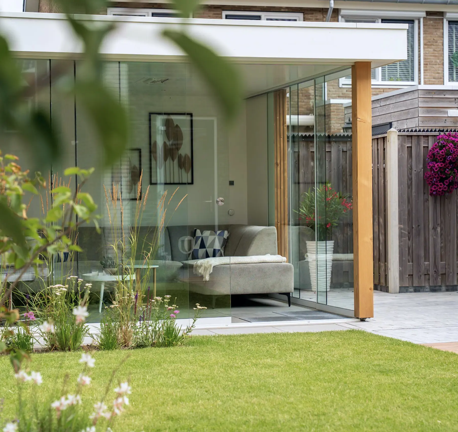 Moderne veranda met glazen wanden en een grijze bank, aangelegd met een groen gazon en kleurrijke bloemen.