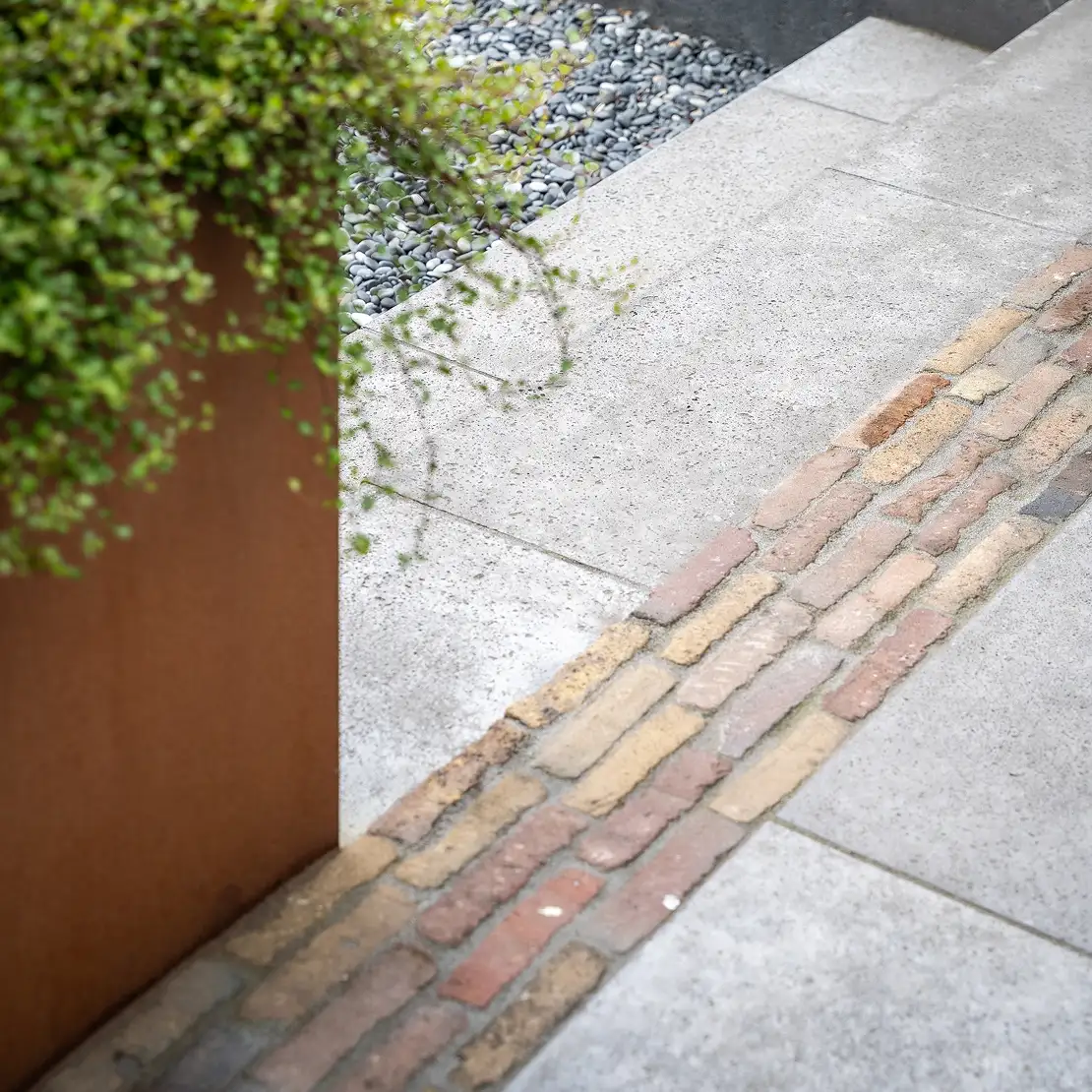 Een geplaveid pad met een strook van oude stenen naast een plantenbak met groen bladerdek en grijze keien achter.
