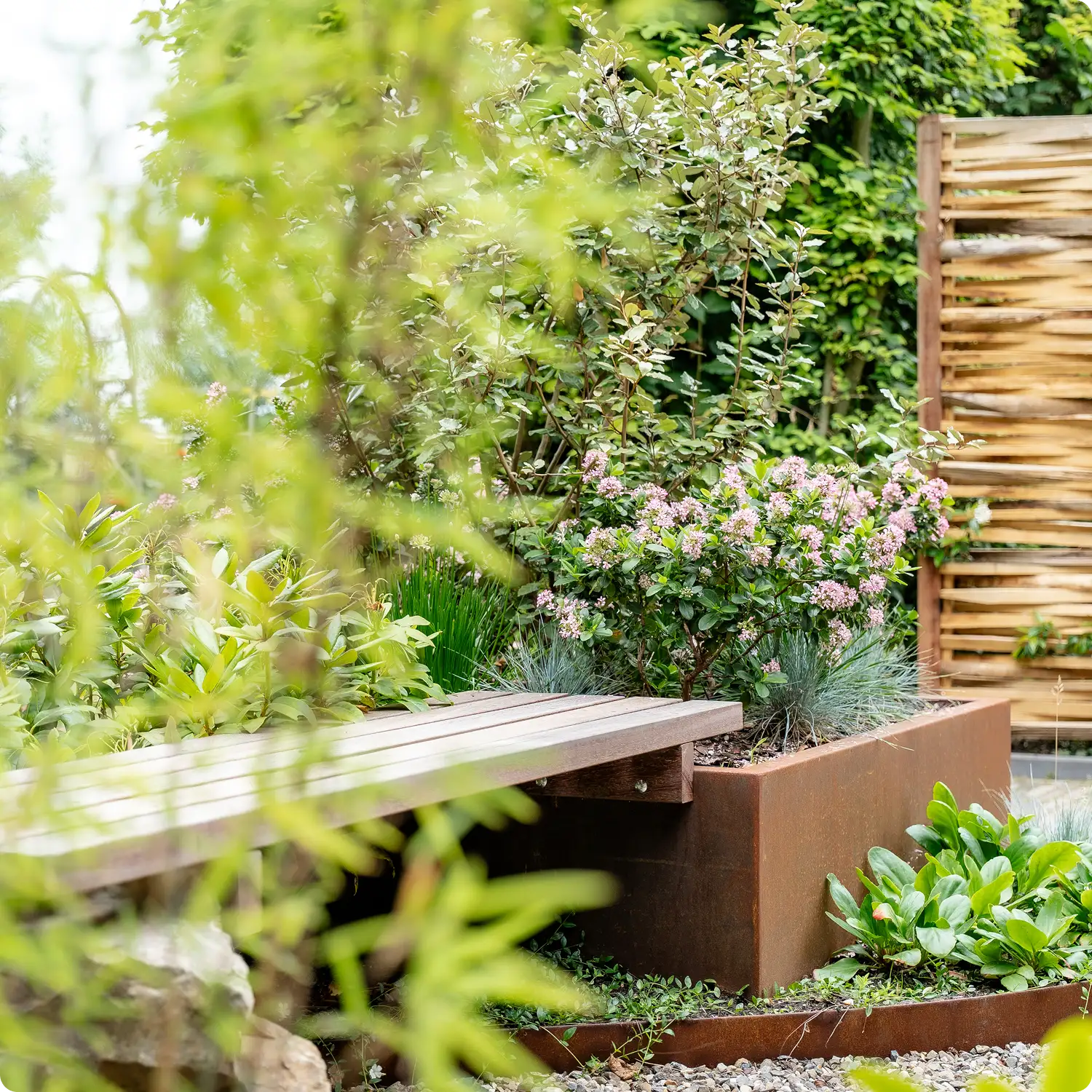 Wooden bench integrated into a garden planter filled with green plants and pink flowers, surrounded by greenery and a wooden privacy screen.