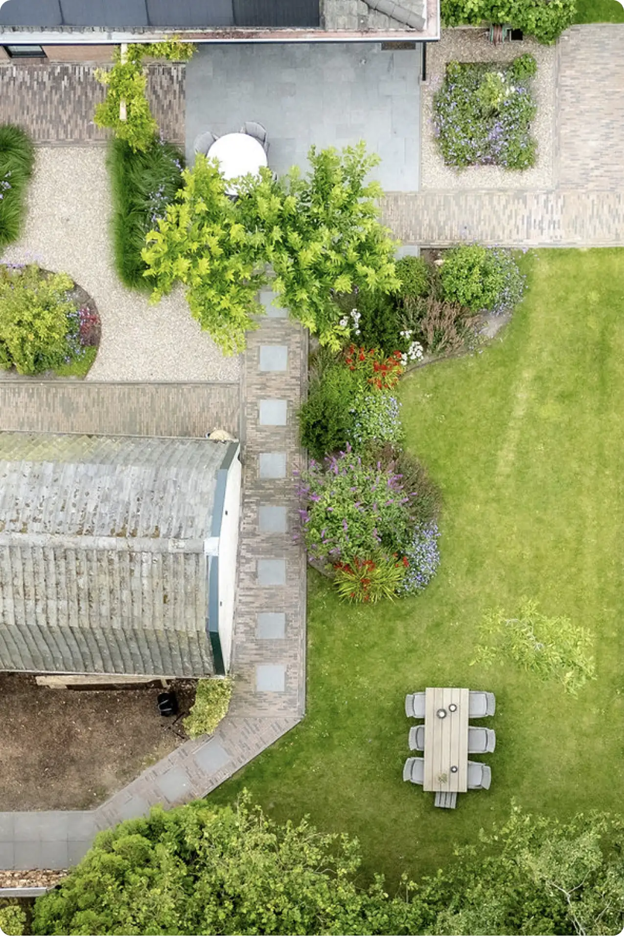Luchtfoto van een tuin met een houten eettafel met zes stoelen op gras, omringd door bloemen, struiken en bomen, naast een huis met een grijs dak en een betegeld terras.