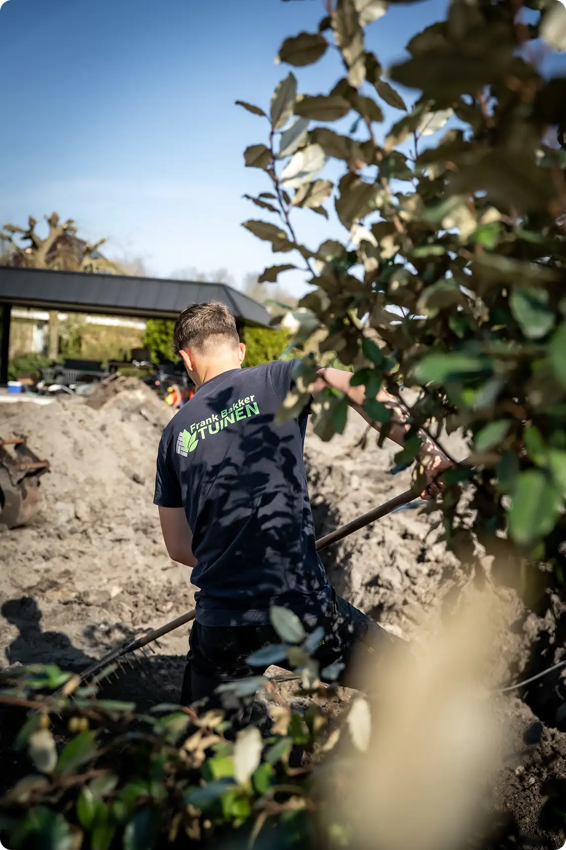 Persoon in een blauw T-shirt met 'Frank Bakker Tuinen' op de rug, werkt buiten met een schop in een tuin met aarde en planten.