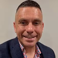 Smiling man with short hair wearing a dark blazer and a checkered shirt against a plain background.