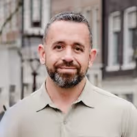 Smiling man with short hair and beard wearing a light green collared shirt standing outdoors in an urban setting.