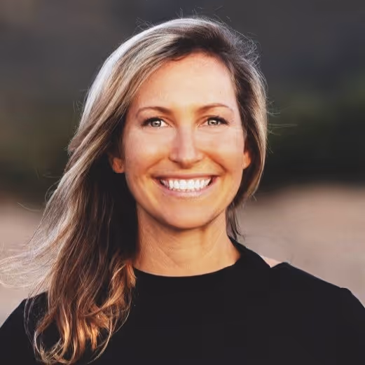 Smiling woman with long light brown hair wearing a black top against a blurred outdoor background.