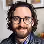 Man with curly hair, glasses, and a beard smiling against a dark background.