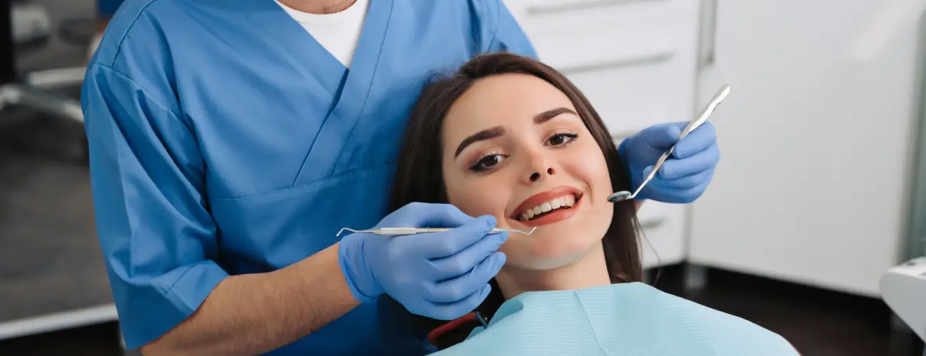 A dentist in blue scrubs holding dental instruments beside a patient in a chair.