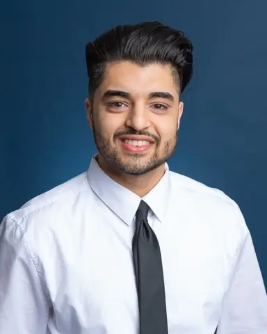 Man in white shirt and black tie with slicked-back hair against a blue background.