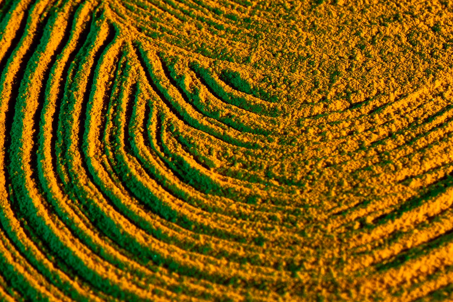 Close-up view of concentric circular patterns in golden sand with green highlights creating a textured, wave-like surface.