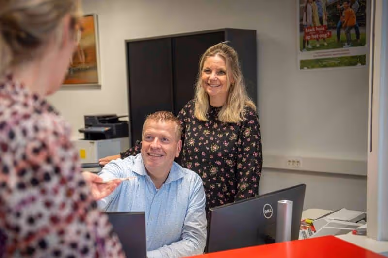 Een man en een vrouw glimlachen terwijl ze achter een balie met computer zitten, kijkend naar een andere vrouw voor hen.