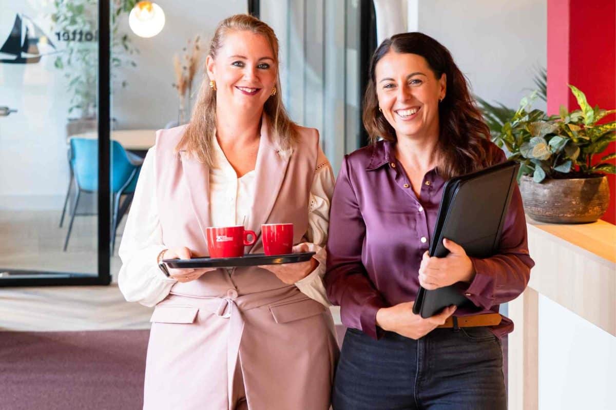 Twee vrouwen glimlachend in een kantoor; één houdt een dienblad met twee rode koffiekopjes, de ander een zwarte map.