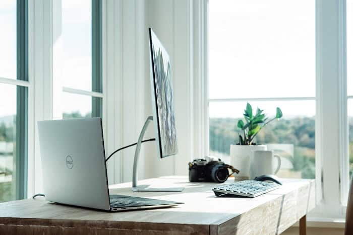 Een hoek van een bureau met een gesloten Dell-laptop, een computerscherm, toetsenbord, muis en een camera bij een raam met uitzicht op bomen.