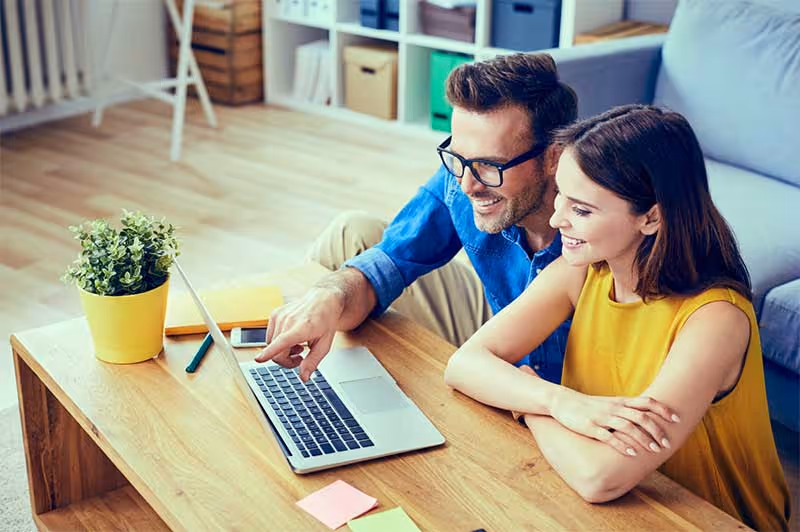 Een lachend stel zit samen aan een houten tafel thuis en kijkt op een laptop.
