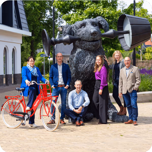 Zes mensen poseren buiten rond een groot zwart beeldenhond die een kettlebell in zijn bek houdt, met een rode fiets op de voorgrond.