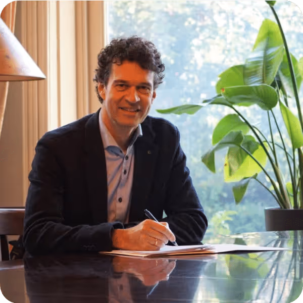 Man met krullend haar en donker colbert zittend aan een tafel, schrijvend in een notitieboekje met een pen, met een grote groene plant naast hem bij een raam.