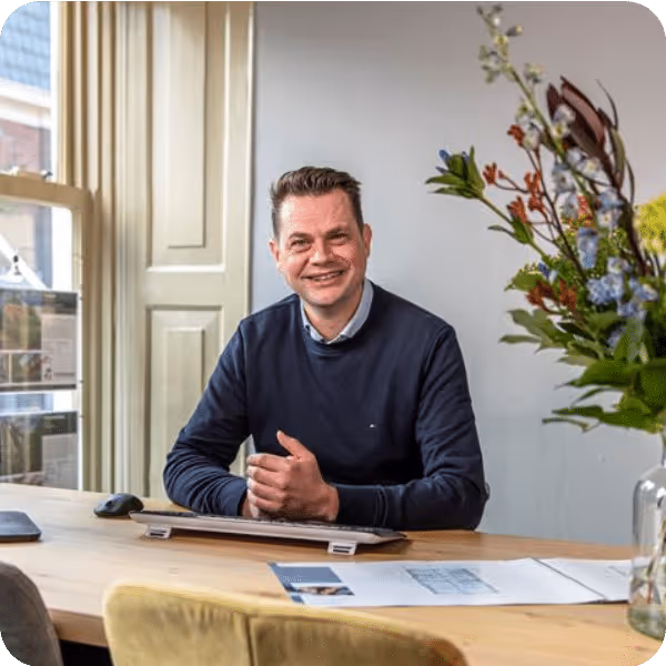 Man in een donkerblauwe trui zittend aan een houten tafel in een lichte kamer met een toetsenbord en papieren, naast een vaas met bloemen.
