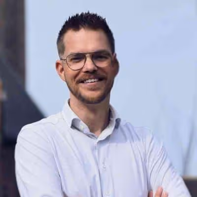 Man met kort donker haar en baard, glimlachend met een witte overhemd en bril, met armen over elkaar.