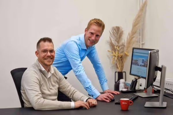 Twee mannen, een zittend en een leunend over een bureau, glimlachen in een moderne kantoorruimte met een computerscherm en een rode koffiekop.