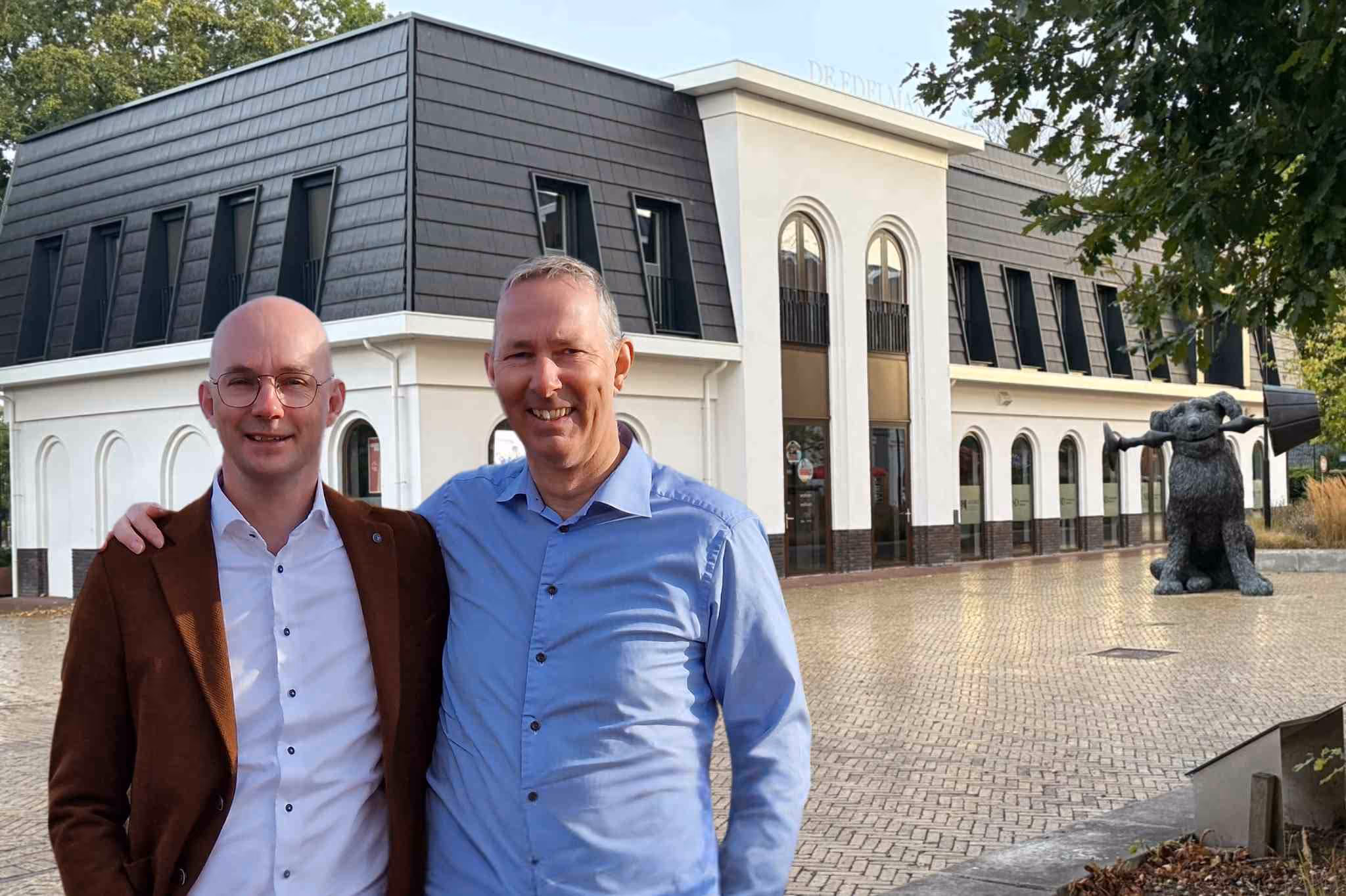 Twee lachende mannen met een arm om elkaars schouder staan voor een modern wit en zwart gebouw met een grote hondensculptuur aan de rechterkant.