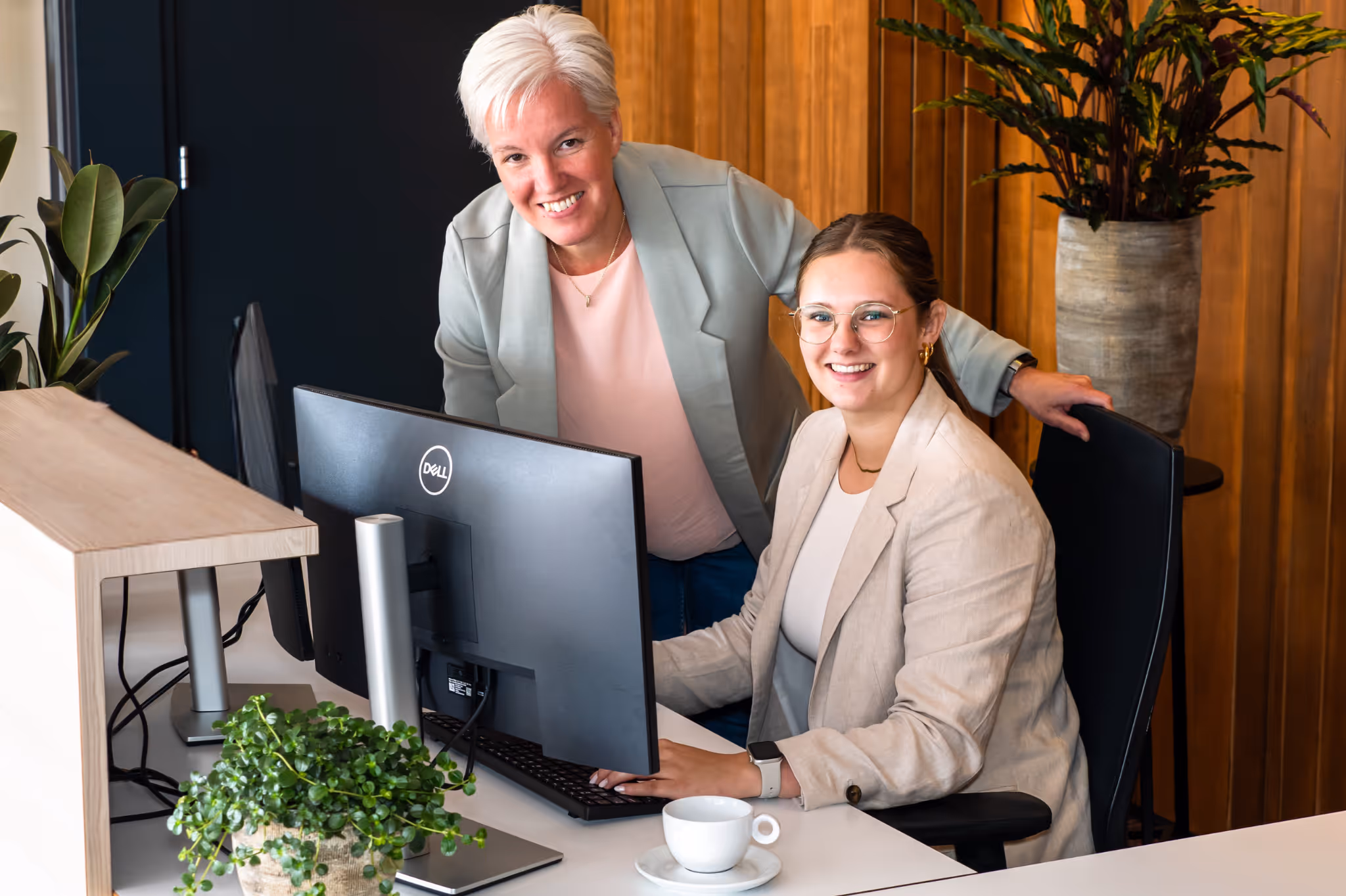 Twee vrouwelijk kantoormedewerkers, één zittend aan een computer en één staand ernaast, glimlachend in een moderne kantooromgeving.