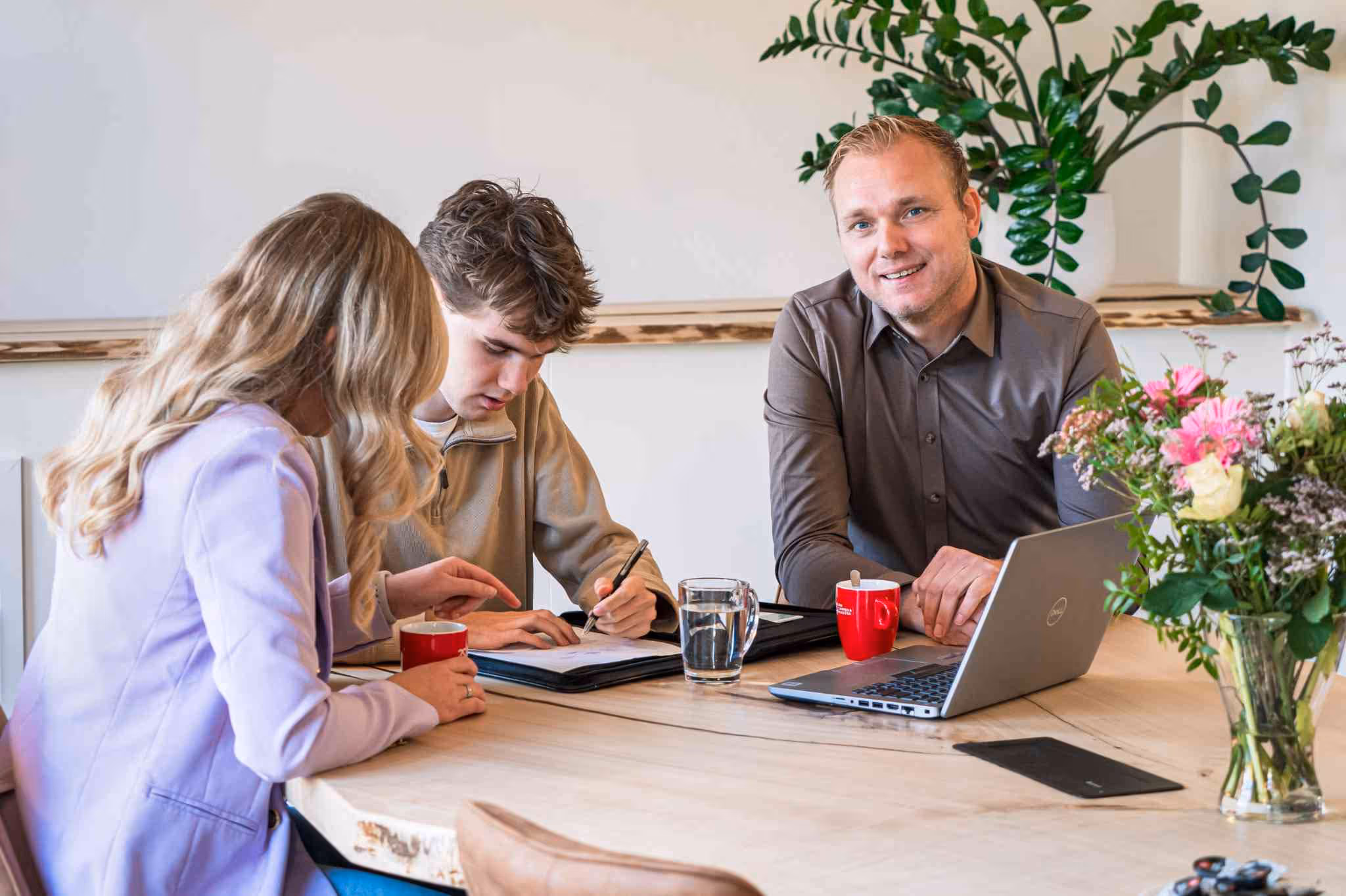 Drie mensen overleggen aan een tafel; een man kijkt glimlachend naar de camera terwijl twee andere personen documenten doornemen en aantekeningen maken.