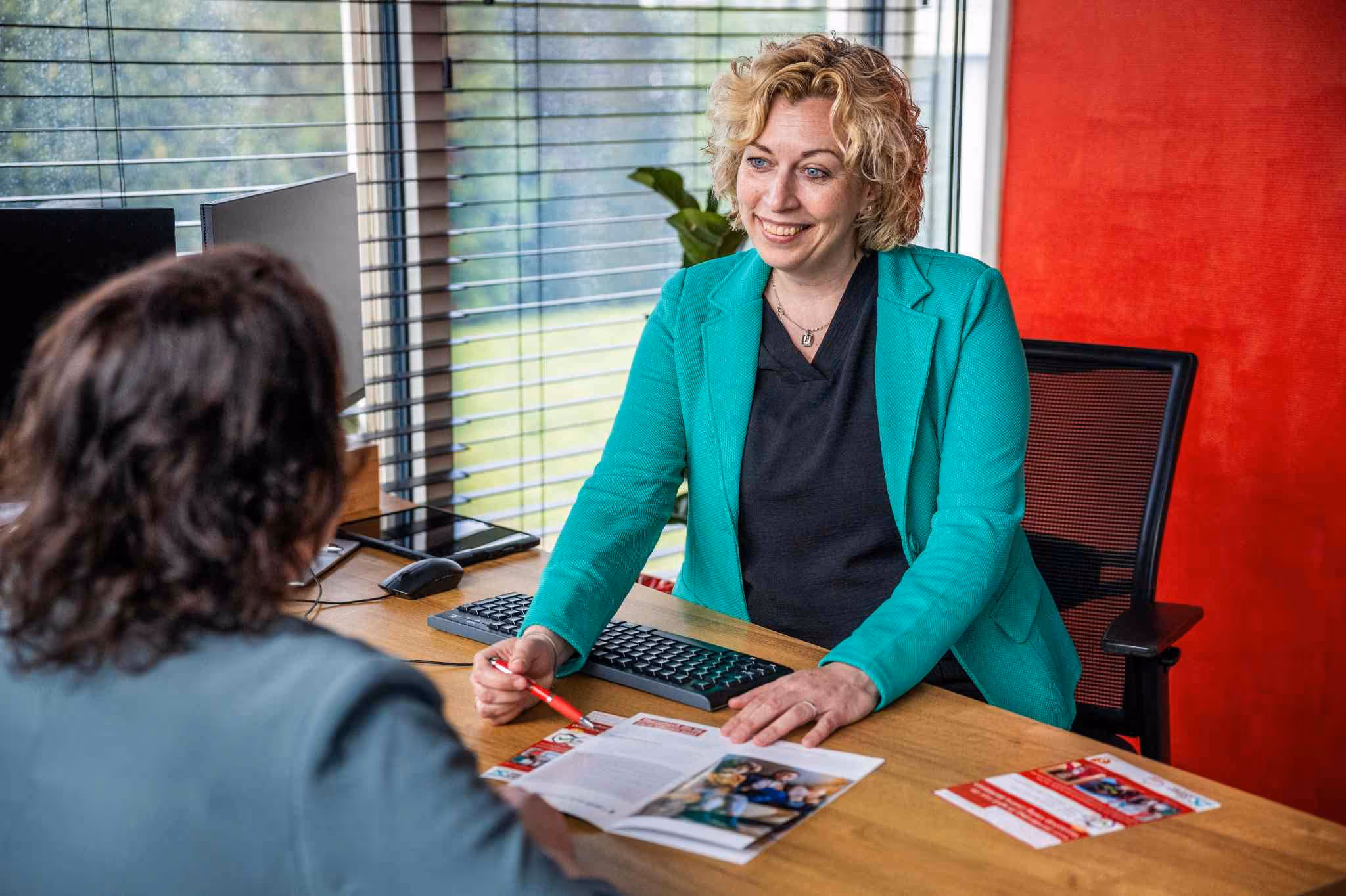 Vrouw met blond krullend haar en een groene blazer die een folder bespreekt met een andere vrouw aan een bureau met computer en rood interieur.