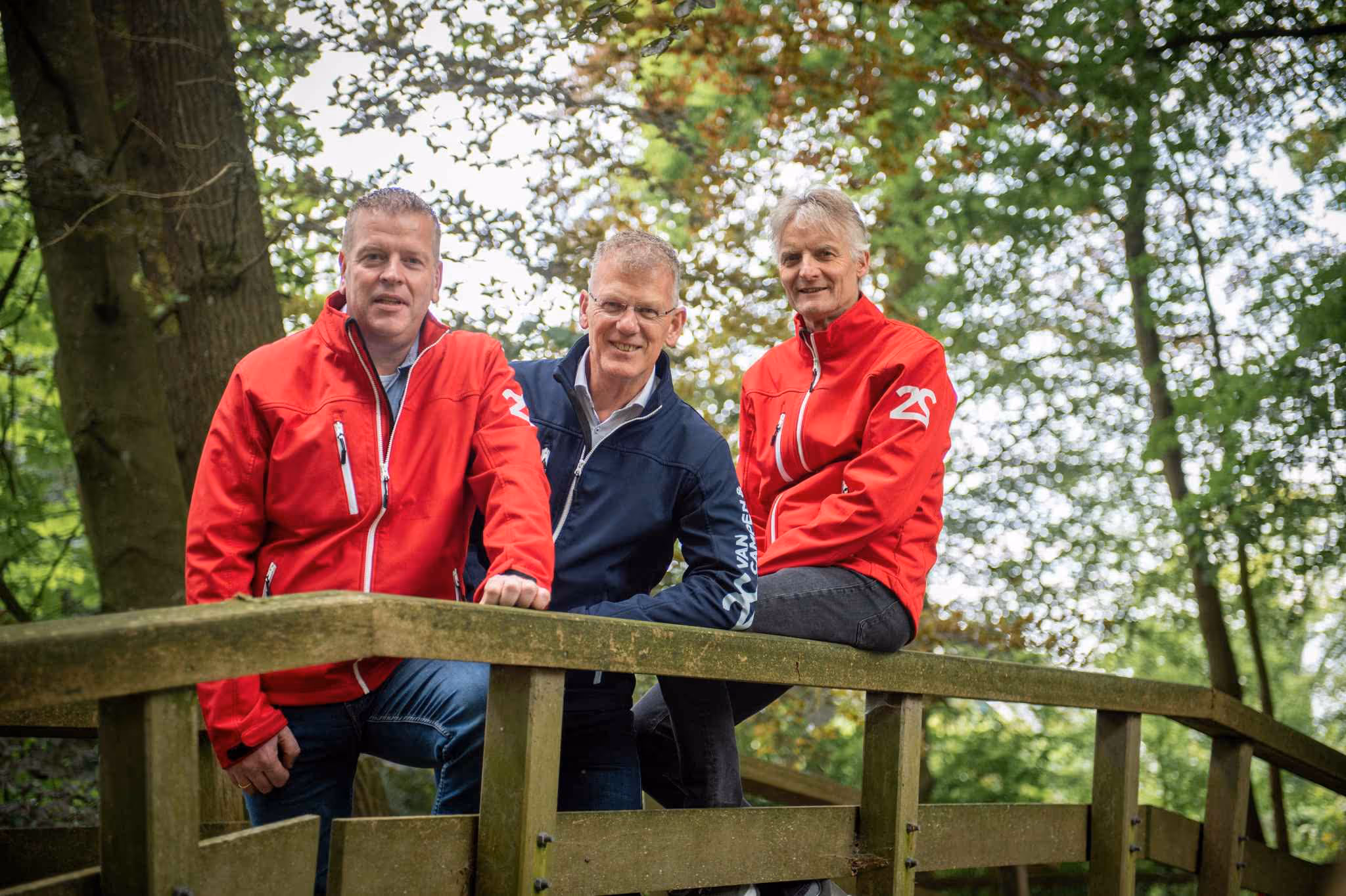 Drie mannen, twee in rode jassen en één in een blauwe jas, poseren op een houten brug in een groene bosrijke omgeving.