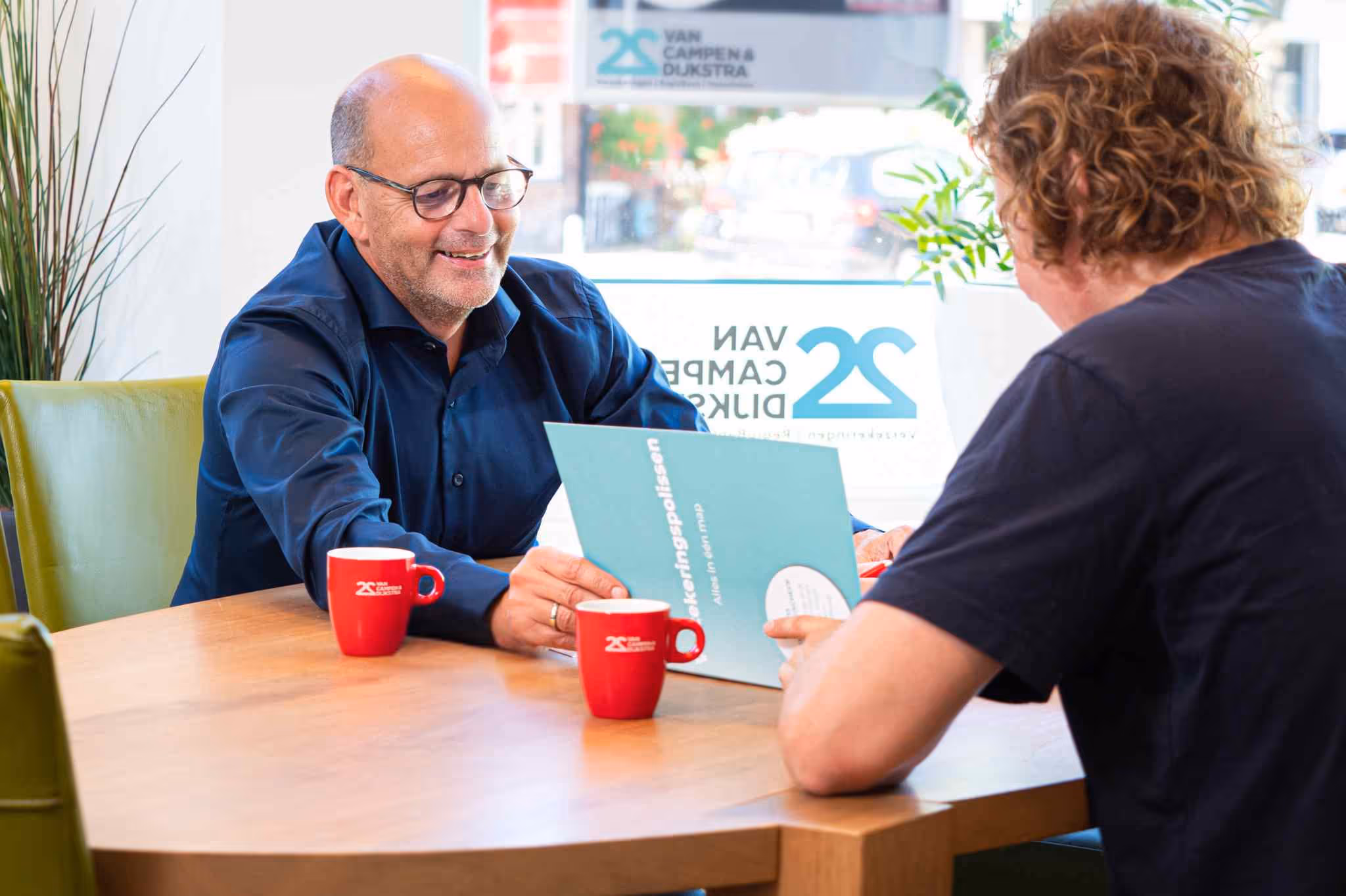 Twee mannen zitten aan een tafel en bespreken een verzekeringsbrochure bij Van Campen & Dijkstra.