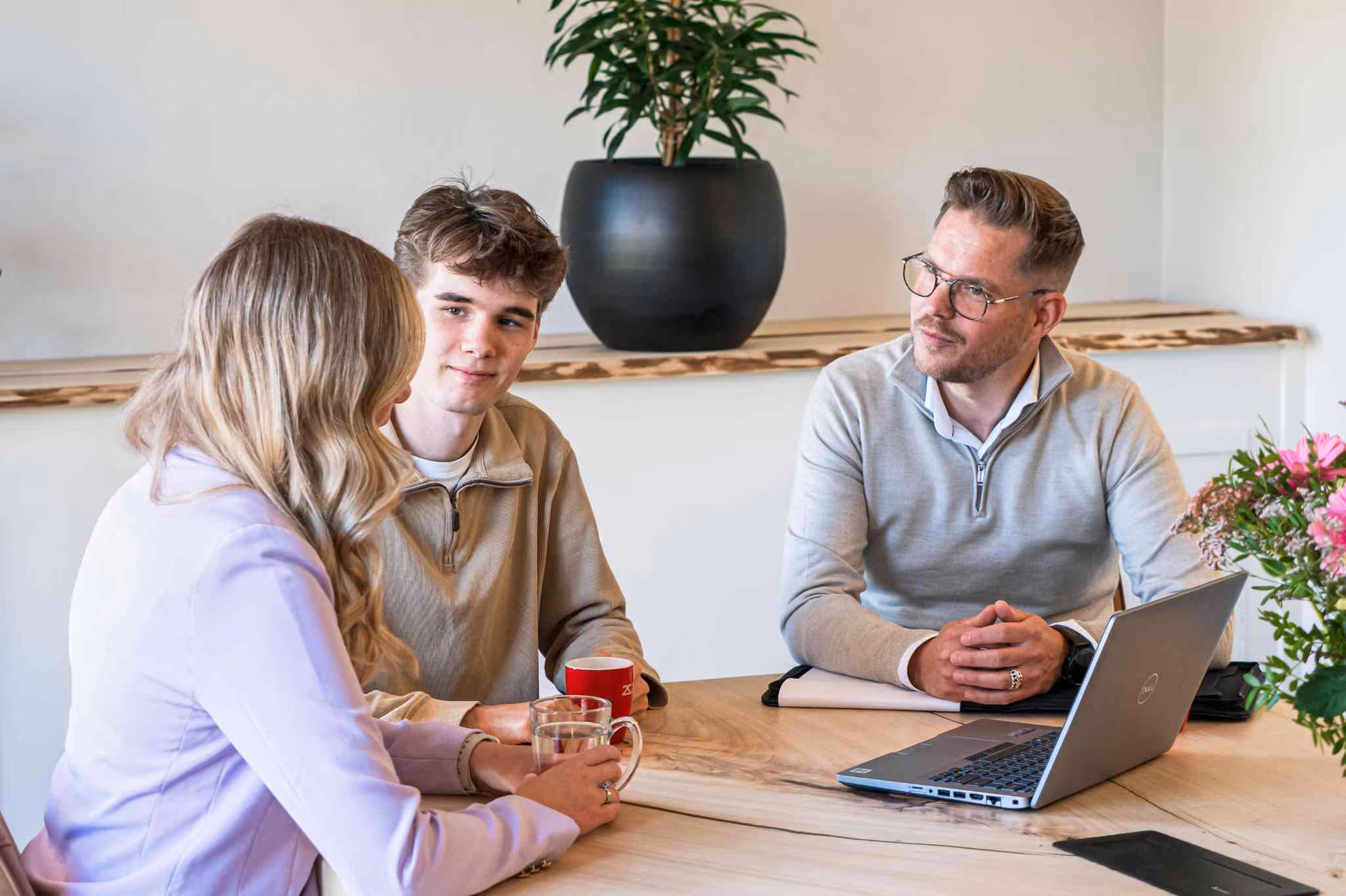 Drie mensen in een informeel overleg aan een houten tafel met een laptop, koffiekop en glas water.