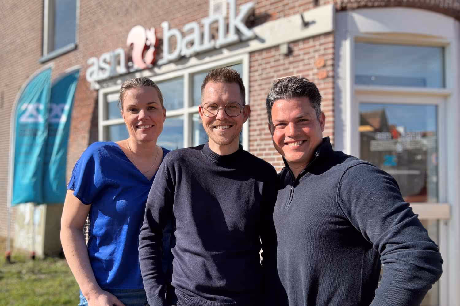 Drie glimlachende volwassenen staan voor een gebouw met het bord 'asn bank'.