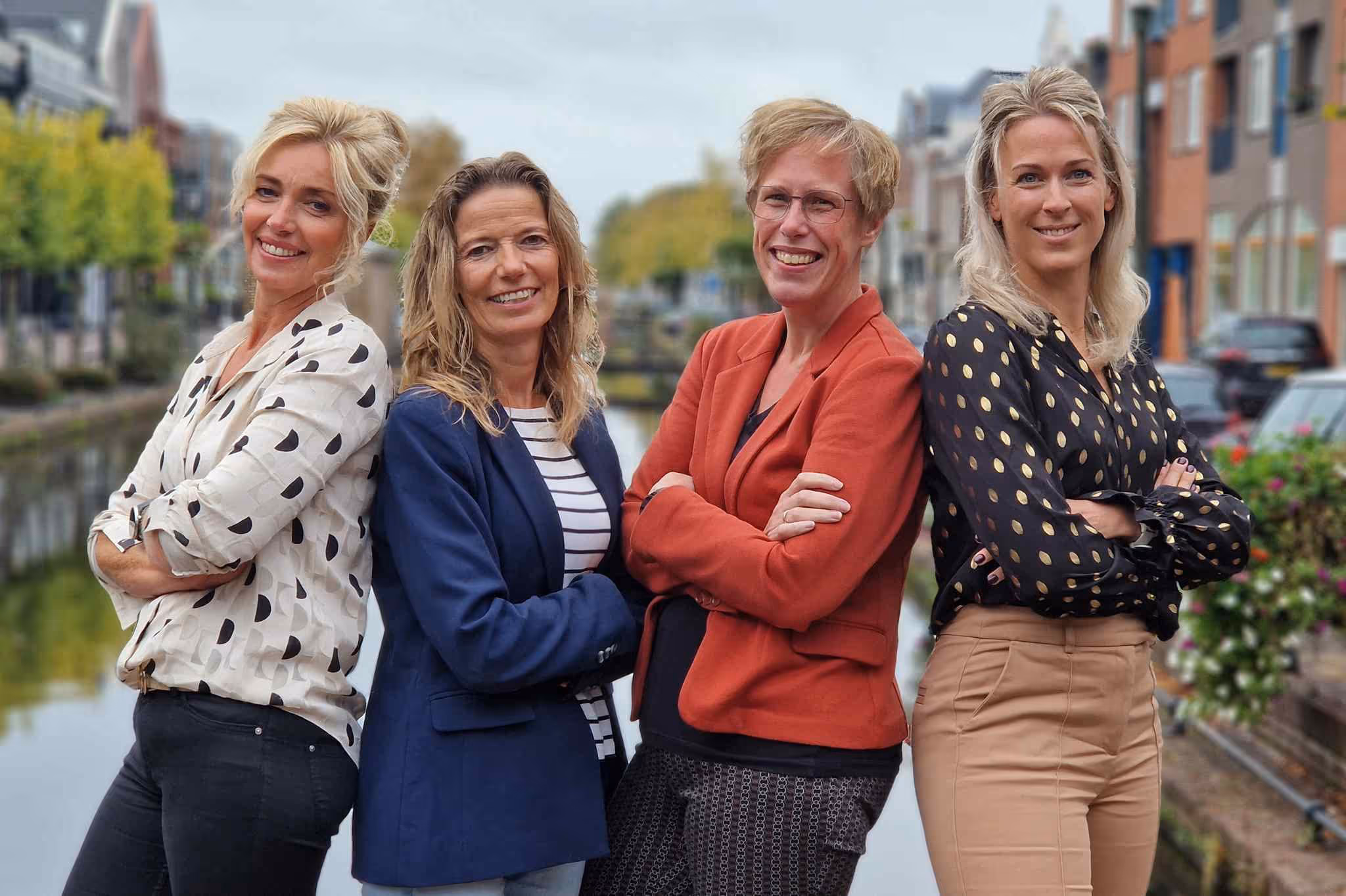 Vier lachende vrouwen met armen over elkaar, staand buiten langs een waterkanaal in een stadsomgeving.