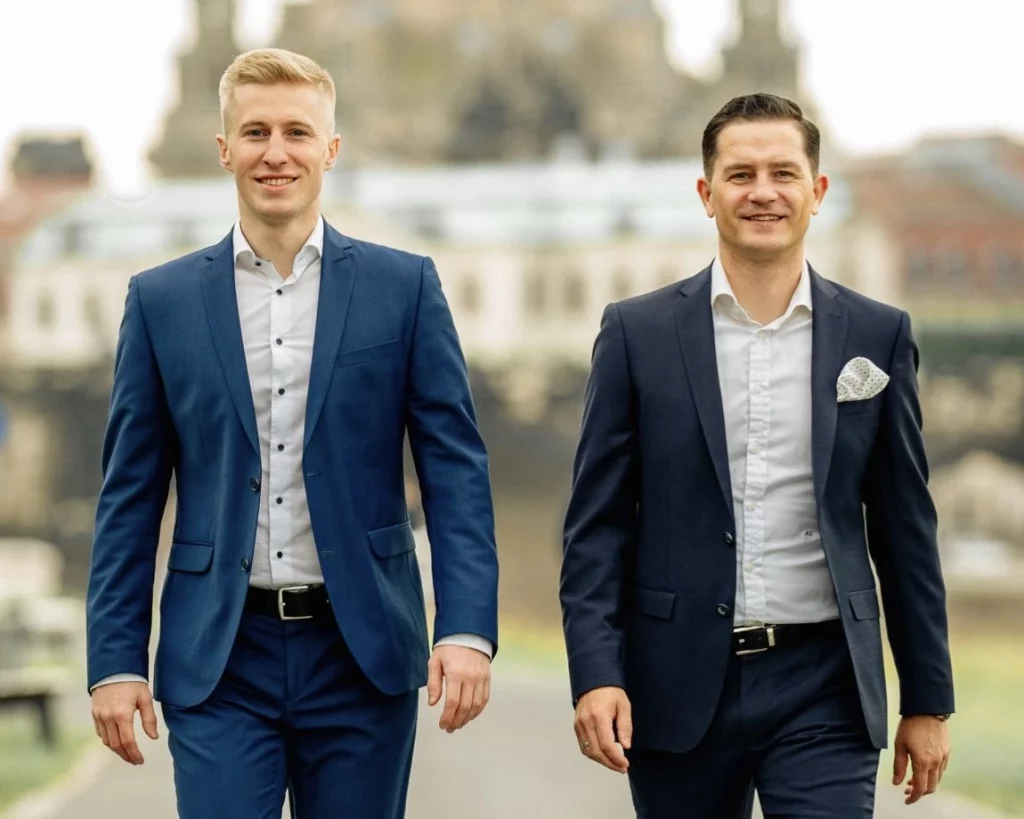Manu und Willi, Gründer von Clear Finance, spazieren selbstbewusst im Anzug durch eine Stadtlandschaft.