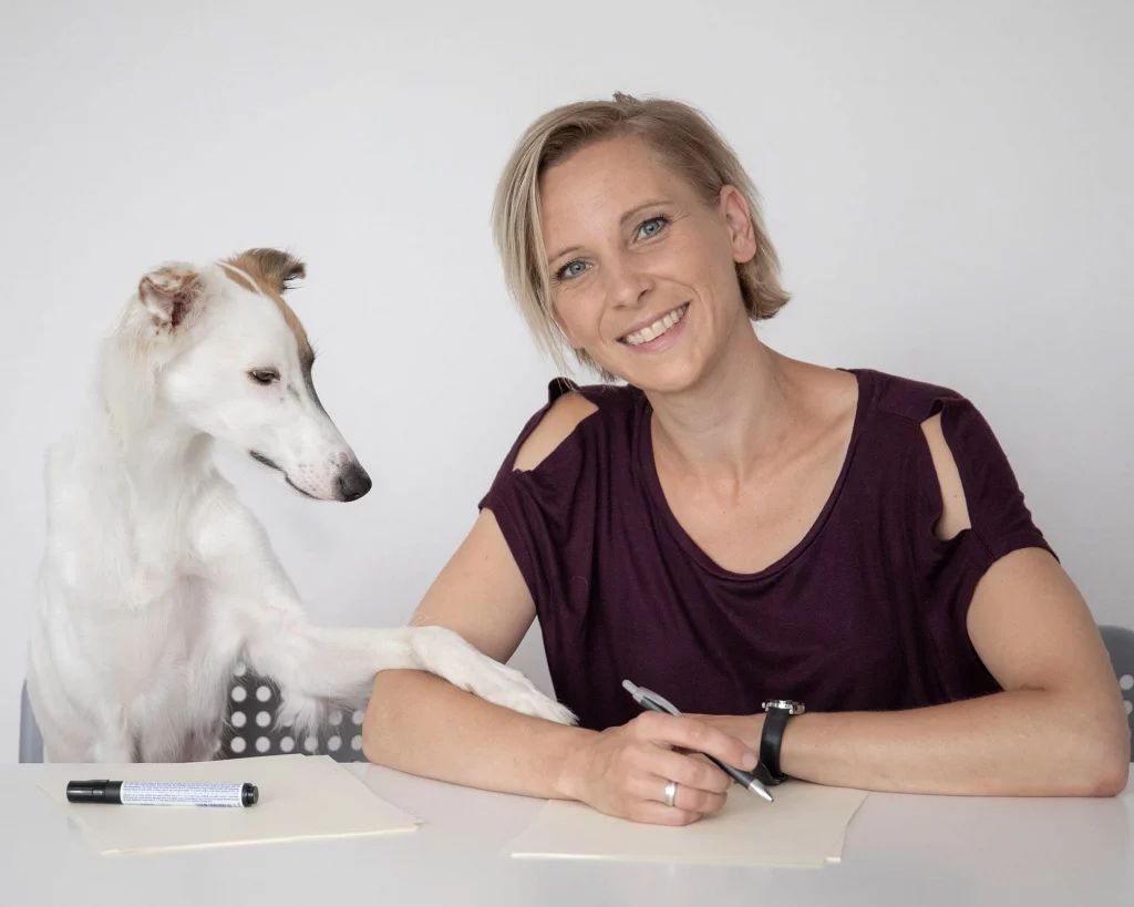 Julia Hammerschmidt, Kundin der Provenia, sitzt lächelnd an einem Tisch mit einem weißen Hund, der eine Pfote auf ihren Arm legt.